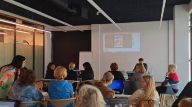 Grupa osób siedzi w nowoczesnej klasie, uczestnicząc w warsztacie. Używają laptopów, a prezentacja jest wyświetlana na ekranie z przodu sali. Atmosfera jest skupiona i nastawiona na współpracę.