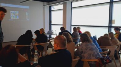 Grupa osób siedzi w klasie, naprzeciwko ekranu projekcyjnego wyświetlającego kolorowe obrazy. Osoba stoi blisko ekranu, angażując publiczność. W pomieszczeniu znajdują się duże okna, wpuszczające naturalne światło.