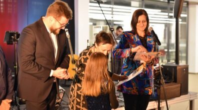 Prezydent miasta, w okularach i eleganckim garniturze wraz z dyrektorką Miejskiej Biblioteki i kierownikiem działu Animacji i Promocji wręczają laureatom dyplomy i nagrody. W tle widoczne są instrumenty muzyczne, mikrofon oraz czerwona i niebieska poświata, co nadaje wydarzeniu formalno-artystyczny charakter. Wnętrze jest przestronne, z przeszklonymi ścianami i nowoczesnym wystrojem.