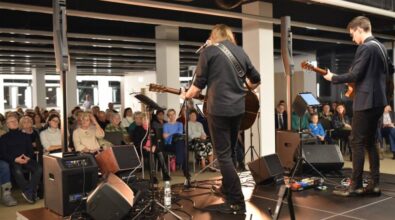 Widok z perspektywy sceny na muzyków oraz publiczność. Dwóch gitarzystów widocznych od tyłu wykonuje utwór na scenie. Jeden z nich trzyma akustyczną gitarę i ma dłuższe włosy, drugi gra na elektrycznej gitarze. Przed nimi stoją mikrofony, monitory odsłuchowe oraz pedały efektów. W tle widać siedzącą publiczność, która uważnie obserwują występ. Sala jest przestronna i nowoczesna, z czarnym sufitem i jasnym oświetleniem. Po prawej stronie ustawione są głośniki i oświetlenie sceniczne.