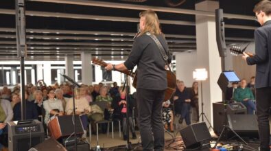 Widok na muzyków oraz publiczność. Dwóch gitarzystów widocznych od tyłu wykonuje utwór na scenie. Przed nimi stoją mikrofony, monitory odsłuchowe oraz pedały efektów. W tle widać siedzącą publiczność.