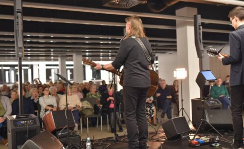 Widok na muzyków oraz publiczność. Dwóch gitarzystów widocznych od tyłu wykonuje utwór na scenie. Przed nimi stoją mikrofony, monitory odsłuchowe oraz pedały efektów. W tle widać siedzącą publiczność.