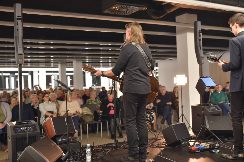 Widok na muzyków oraz publiczność. Dwóch gitarzystów widocznych od tyłu wykonuje utwór na scenie. Przed nimi stoją mikrofony, monitory odsłuchowe oraz pedały efektów. W tle widać siedzącą publiczność.