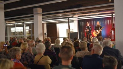 Na scenie występuje duet muzyków grających na gitarach, oświetlony światłami w odcieniach czerwieni i niebieskiego. Publiczność siedzi na krzesłach w rzędach, uważnie słuchając występu.