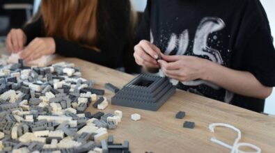 Dwie osoby budują konstrukcje z małych klocków w odcieniach szarości i bieli. Na drewnianym stole znajduje się wiele rozsypanych elementów, a jedna z osób skupia się na składaniu niewielkiej piramidy z klocków.