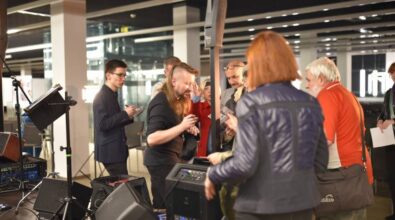 Muzycy stoją przy stanowisku z nagłośnieniem i sprzętem technicznym i podpisują płyty. Obok stoją uczestnicy wydarzenia, którzy czekają na swoją kolej.