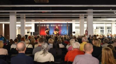 Na scenie występuje duet muzyków grających na gitarach, oświetlony światłami w odcieniach czerwieni i niebieskiego. Publiczność siedzi na krzesłach w rzędach, uważnie słuchając występu. W tle widoczne są regały z książkami oraz nowoczesne wnętrze.