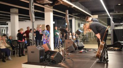 Muzycy kłaniają się, a widownia bije brawo. Część osób stoi, część siedzi. Kobieta w kolorowej marynarce przemawia do uczestników wydarzenia.