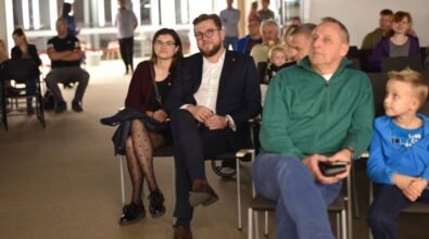 Na zdjęciu widzimy uczestników wydarzenia siedzących na krzesłach w sali. Na pierwszym planie znajduje się para, kobieta w eleganckiej spódnicy i marynarce oraz mężczyzna w garniturze, oboje uśmiechnięci i zwróceni w stronę fotografa. Jest to prezydent miasta razem z małżonką. Obok nich siedzą inni uczestnicy. W tle widoczne są kolejne osoby oraz przestrzeń sali, z nowoczesnym oświetleniem i dużymi oknami.