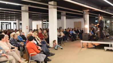 Na zdjęciu widzimy dużą grupę osób uczestniczących w wydarzeniu, w przestronnej sali. Widownia składa się głównie z osób dorosłych i seniorów, a w pierwszym rzędzie siedzą również dzieci. Po prawej stronie znajduje się scena, na której występują artyści z gitarami, skierowani w stronę publiczności.