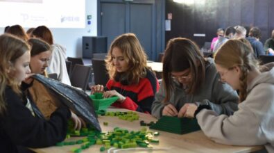 Grupa młodych osób siedzi przy stole i skupia się na tworzeniu konstrukcji z zielonych klocków. Na pierwszym planie widoczne są cztery dziewczyny w różnym wieku, które starannie układają elementy. W tle widać inne osoby biorące udział w warsztatach w jasnym, przestronnym pomieszczeniu. Na ekranie w tle widoczne są logotypy organizatorów: Piotrkowa Trybunalskiego i Biblioteki.