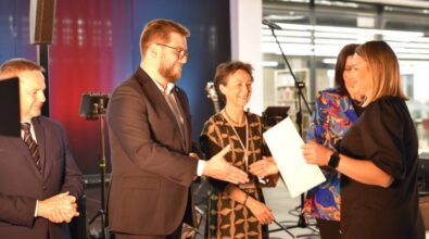 Prezydent miasta wraz z dyrektorką Miejskiej Biblioteki i kierownikiem działu Animacji i Promocji wręczają laureatom dyplomy i nagrody. W tle widoczne są instrumenty muzyczne, mikrofon oraz czerwona i niebieska poświata, co nadaje wydarzeniu formalno-artystyczny charakter. Wnętrze jest przestronne, z przeszklonymi ścianami i nowoczesnym wystrojem.