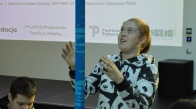 Chłopiec asekuruje wysoki model zbudowany z niebieskich klocków, podczas gdy inne dzieci pracują przy stołach.