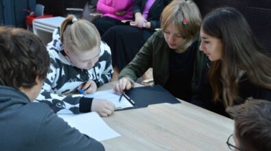 Grupa młodych osób pracuje wspólnie przy stole, skupiając się na zadaniu pisemnym. Jedna z osób trzyma długopis, wskazując coś na kartce, a pozostali uważnie obserwują. W tle widać osoby siedzące na krzesłach, przyglądające się wydarzeniu.