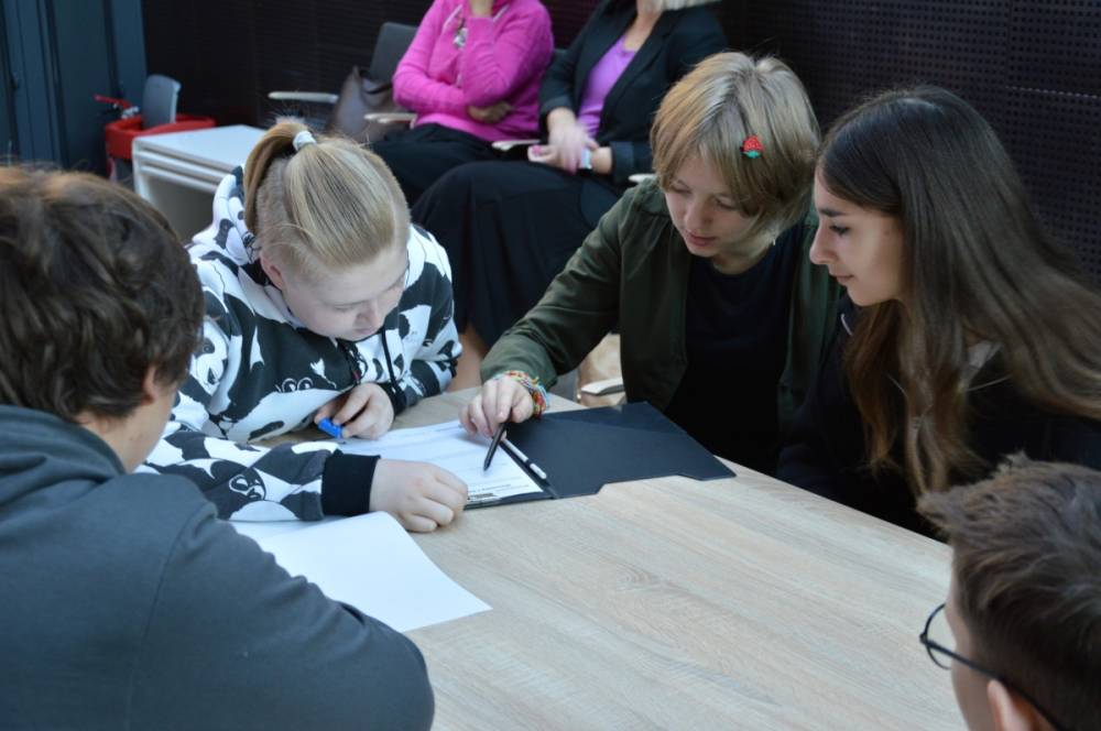 Grupa młodych osób pracuje wspólnie przy stole, skupiając się na zadaniu pisemnym. Jedna z osób trzyma długopis, wskazując coś na kartce, a pozostali uważnie obserwują. W tle widać osoby siedzące na krzesłach, przyglądające się wydarzeniu.