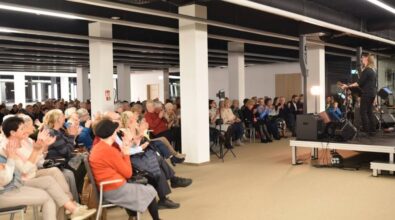 Na zdjęciu widzimy dużą grupę osób uczestniczących w wydarzeniu, w przestronnej sali. Widownia składa się głównie z osób dorosłych i seniorów, a w pierwszym rzędzie siedzą również dzieci. Po prawej stronie znajduje się scena, na której występują artyści z gitarami, skierowani w stronę publiczności.