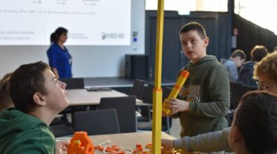 Chłopiec trzyma wysoki model zbudowany z żółtych klocków, podczas gdy inne dzieci pracują przy stołach. W tle widoczna jest prowadząca warsztaty na tle slajdu wyświetlanego na ekranie.