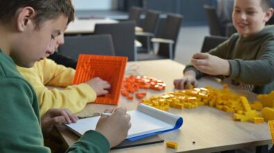 Przy stoliku siedzą uczestnicy, którzy budują konstrukcje z żółtych i pomarańczowych klocków. Na stole znajduje się wiele rozsypanych elementów, jedna z osób robi jakieś zapiski, a druga buduje pomarańczową piramidę.