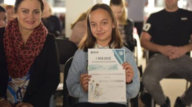 Uśmiechnięta dziewczynka trzymająca w rękach dyplom za zajęcie pierwszego miejsca w konkursie plastycznym 'Mediateka na zakładce'. Obok niej siedzi kobieta w szalu w panterkę. W tle widać osoby siedzące na krzesłach, w rozmytym tle sali.