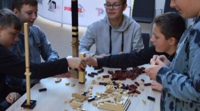 Kolejna grupa uczestników buduje wysoki model z czarnych, brązowych i beżowych klocków. W tle widoczna ścianka reklamująca bibliotekę i kino Piksel.