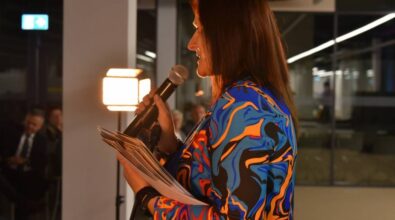 Kierownik działu Animacji i Promocji trzyma mikrofon w jednej ręce oraz plik kartek w drugiej, przemawia na stojąco. Ubrana jest w kolorową marynarkę o wzorze przypominającym abstrakcyjne linie w żywych barwach, takich jak niebieski, pomarańczowy i czarny. Zdjęcie wykonano z boku, ukazując ją w półprofilu na tle nowoczesnego wnętrza z przeszklonymi ścianami. Po lewej stronie w tle widoczne są osoby siedzące.