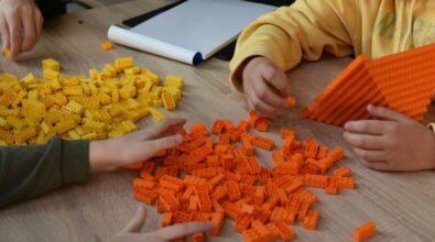 Przy stoliku siedzą uczestnicy, którzy budują konstrukcje z żółtych i pomarańczowych klocków. Jedna z osób buduje piramidę w pomarańczowym kolorze. Druga ma przed sobą szkicownik.