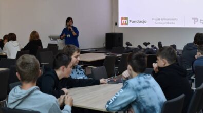 Uczestnicy siedzią przy stolikach i słuchają osobę prowadzącą warsztaty. Na ekranie w tle widoczne są logotypy organizatorów: Piotrkowa Trybunalskiego Fundacji mBank.