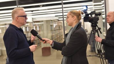 Na zdjęciu widzimy wywiad przeprowadzany w nowoczesnym wnętrzu, prawdopodobnie w bibliotece lub innym miejscu z regałami książkowymi. Mężczyzna w ciemnym ubraniu i okularach odpowiada na pytania, trzymając w dłoni długopis, podczas gdy kobieta w płaszczu i szaliku trzyma mikrofon skierowany w jego stronę. W tle stoi operator kamery z profesjonalnym sprzętem, rejestrujący rozmowę. Światło odbija się od szklanych powierzchni, tworząc jasną, profesjonalną atmosferę.