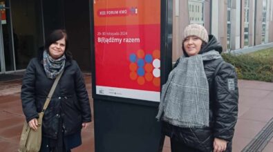 dwie uśmiechnięte osoby stojące obok tablicy informacyjnej promującej XIII Forum KMO (Klubu Młodego Odkrywcy). Tablica zawiera informacje o wydarzeniu, które odbywa się w dniach 29–30 listopada 2024 roku pod hasłem „BĄDŹMY RAZEM”. W tle widoczny jest nowoczesny budynek, prawdopodobnie miejsce, gdzie odbywa się forum. Osoby na zdjęciu są ciepło ubrane, co sugeruje chłodniejszą porę roku