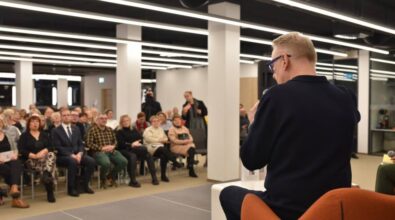 Na zdjęciu widzimy widownię zgromadzoną na spotkaniu. Ludzie siedzą na krzesłach w dobrze oświetlonej sali, z regularnym układem białych słupów i sufitowych paneli świetlnych. Na pierwszym planie, po prawej stronie, z tyłu widać osobę siedzącą na pomarańczowym fotelu, trzymającą mikrofon. W tle znajduje się kamera na statywie, a publiczność uważnie słucha.