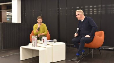 Na zdjęciu widzimy dwie osoby siedzące na pomarańczowych fotelach na scenie, podczas rozmowy w trakcie wydarzenia. Osoba po lewej stronie, ubrana w żółty sweter, trzyma mikrofon i wygląda na uśmiechniętą, podczas gdy osoba po prawej, w ciemnym stroju, również trzyma mikrofon i uśmiecha się w stronę swojego rozmówcy. Między nimi na stoliku znajdują się książki i szklanki z wodą. Tło sceny jest czarne, a atmosfera zdjęcia jest swobodna i skupiona na rozmowie. Wydarzenie ma miejsce w nowoczesnej bibliotece.