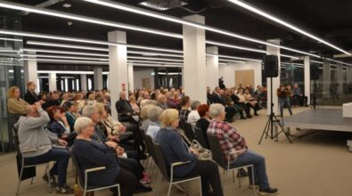 Na zdjęciu widoczna jest duża grupa osób uczestniczących w spotkaniu. Przestrzeń jest jasna, nowoczesna, z charakterystycznym oświetleniem sufitowym w postaci długich, prostokątnych lamp. W tle znajduje się scena wyposażona w sprzęt nagłaśniający. Uczestnicy siedzą na krzesłach, uważnie słuchając. Widoczna jest różnorodność wiekowa, choć dominują osoby starsze. W tle znajdują się przeszklone drzwi oraz część osób stojących. Atmosfera wydaje się skupiona, co sugeruje udział w wydarzeniu kulturalnym.