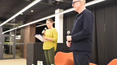 Na zdjęciu widzimy dwie osoby stojące na scenie podczas wydarzenia literackiego. Mężczyzna w granatowym stroju, trzymający mikrofon, stoi z boku, obserwując sytuację. Obok niego znajduje się kobieta ubrana w żółty sweter i zielone spodnie, również z mikrofonem w dłoni, czytająca tekst z notatek na klipbordzie. W tle widoczne są ciemne zasłony i nowoczesne oświetlenie, a na pierwszym planie fragment stolika z książkami i szklanką wody. Atmosfera sugeruje formalne, ale przyjazne spotkanie autorskie lub dyskusję literacką.