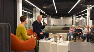 Na zdjęciu widać dwie osoby siedzące na pomarańczowych fotelach na scenie podczas wydarzenia. Osoba po lewej stronie, ubrana w zielony sweter, trzyma mikrofon i zwraca się w stronę rozmówcy. Osoba po prawej stronie, w ciemnym stroju, przemawia do mikrofonu, patrząc na publiczność. Na stoliku przed nimi znajdują się książki oraz szklanki z wodą. W tle widać nowoczesne wnętrze z przeszklonymi balustradami, a po prawej stronie fragment publiczności, która uśmiecha się i z uwagą słucha rozmowy.