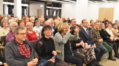 Na zdjęciu widzimy zgromadzoną publiczność uczestniczącą w wydarzeniu literackim. W pierwszym rzędzie siedzi kobieta w okularach, ubrana w ciemną sukienkę z delikatnym wzorem, obok niej inna kobieta robi zdjęcia telefonem komórkowym. Obok nich zasiadają mężczyzna w czerwonej koszuli w kratę oraz kilka osób w eleganckich ubraniach. W tle widać pełną salę z zainteresowanymi uczestnikami, w różnym wieku, z uśmiechami na twarzach. Wystrój sali jest nowoczesny, z jasnym oświetleniem i prostymi ścianami. Atmosfera wskazuje na ciekawe i przyjazne spotkanie.