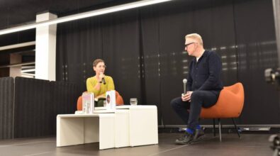 Na zdjęciu widzimy dwie osoby siedzące na pomarańczowych fotelach na scenie, podczas rozmowy w trakcie wydarzenia. Osoba po lewej stronie, ubrana w żółty sweter, trzyma mikrofon, podczas gdy osoba po prawej, w ciemnym stroju, również trzyma mikrofon i patrzy w stronę swojego rozmówcy. Między nimi na stoliku znajdują się książki i szklanki z wodą. Tło sceny jest czarne, a atmosfera zdjęcia jest swobodna i skupiona na rozmowie. Wydarzenie ma miejsce w nowoczesnej sali konferencyjnej lub bibliotece.
