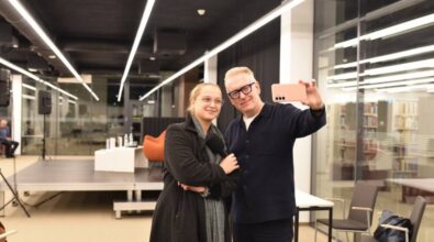 Na zdjęciu widzimy uśmiechniętą kobietę i mężczyznę pozujących do wspólnego selfie w nowoczesnym wnętrzu w bibliotece. Kobieta, ubrana w ciemny płaszcz i szal, trzyma mikrofon, a mężczyzna w okularach i ciemnym swetrze robi zdjęcie telefonem o różowym etui. W tle widać scenę z krzesłami i stolikiem, a także regały z książkami za szklaną ścianą, co tworzy profesjonalną, ale przyjazną atmosferę.