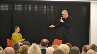 Na zdjęciu widać dwie osoby biorące udział w spotkaniu autorskim. Kobieta w okularach, ubrana w zielony sweter, siedzi na pomarańczowym fotelu i trzyma mikrofon, słuchając z uwagą. Obok niej stoi mężczyzna w ciemnym ubraniu, przemawiający do publiczności z mikrofonem w ręku. W tle znajduje się czarna kotara, a na pierwszym planie widoczne są głowy publiczności, która uważnie śledzi rozmowę. Atmosfera wydaje się być skupiona i profesjonalna.