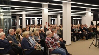Na zdjęciu widoczna jest duża grupa osób uczestniczących w spotkaniu. Przestrzeń jest jasna, nowoczesna, z charakterystycznym oświetleniem sufitowym w postaci długich, prostokątnych lamp. Uczestnicy siedzą na krzesłach, uważnie słuchając. Widoczna jest różnorodność wiekowa, choć dominują osoby starsze. W tle znajdują się przeszklone drzwi oraz część osób stojących. Atmosfera wydaje się skupiona, co sugeruje udział w wydarzeniu kulturalnym.
