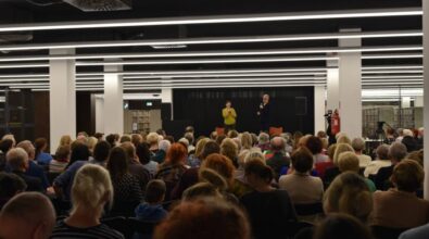 Na zdjęciu widzimy dużą salę wypełnioną publicznością, skierowaną w stronę sceny, na której znajdują się dwie osoby prowadzące wydarzenie. Jedna z osób trzyma mikrofon, a druga dokumenty. Tło sceny jest czarne, a po bokach widoczne są głośniki. Wystrój sali jest nowoczesny, z jasnym oświetleniem w postaci długich, równoległych lamp na suficie. W tle znajdują się regały z książkami, co sugeruje, że wydarzenie odbywa się w bibliotece. Atmosfera zdjęcia wskazuje na formalne, literackie lub kulturalne spotkanie.