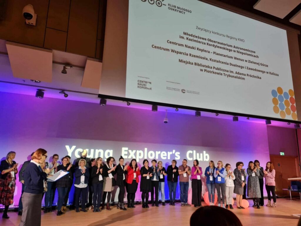 uroczystość rozdania nagród w ramach konkursu "Regiony KMO" organizowanego przez Centrum Nauki Kopernik. Na scenie stoi grupa laureatów i uczestników, którzy otrzymują wyróżnienia. W tle widoczny jest duży ekran z wyświetlonym tekstem informującym o zwycięzcach konkursu. Wyróżnione zostały m.in.: Na scenie widnieje napis „Young Explorer’s Club”. Sala jest nowoczesna, z oświetleniem w odcieniach fioletu, a widownia znajduje się poza kadrem.