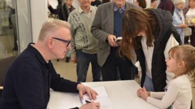 Na zdjęciu widzimy autora siedzącego przy stole, który z pełnym skupieniem podpisuje książkę. Przy stole stoją młoda kobieta pochylająca się w rozmowie z uśmiechniętą dziewczynką, która wygląda na podekscytowaną i zafascynowaną. W tle widać starszego mężczyznę w garniturze i koszuli w kratę, a także inne osoby w kolejce, oczekujące na autograf. Wnętrze biblioteki jest nowoczesne i jasne, co nadaje scenie przyjazną i ciepłą atmosferę.