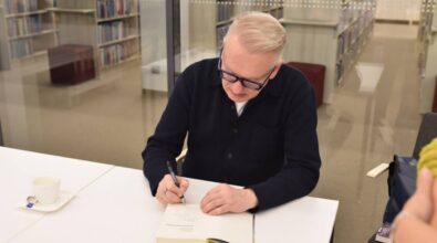 Na zdjęciu widzimy pisarza siedzącego przy białym stole, który z pełnym skupieniem podpisuje książkę. Na stole stoi filiżanka po kawie z łyżeczką na spodku, co sugeruje dłuższą, intensywną pracę podczas spotkania autorskiego. W tle znajduje się szklana ściana, za którą widać regały z książkami w bibliotece. Atmosfera jest spokojna i skupiona, oddając klimat literackiego wydarzenia.