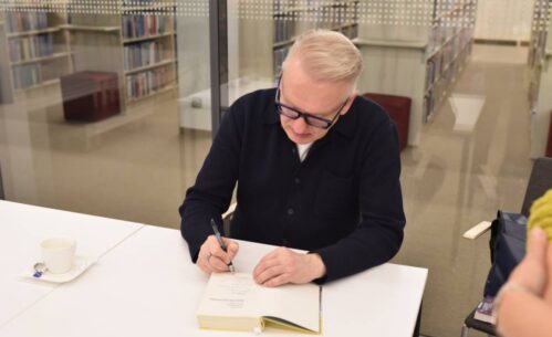 Na zdjęciu widzimy pisarza siedzącego przy białym stole, który z pełnym skupieniem podpisuje książkę. Na stole stoi filiżanka po kawie z łyżeczką na spodku, co sugeruje dłuższą, intensywną pracę podczas spotkania autorskiego. W tle znajduje się szklana ściana, za którą widać regały z książkami w bibliotece. Atmosfera jest spokojna i skupiona, oddając klimat literackiego wydarzenia.