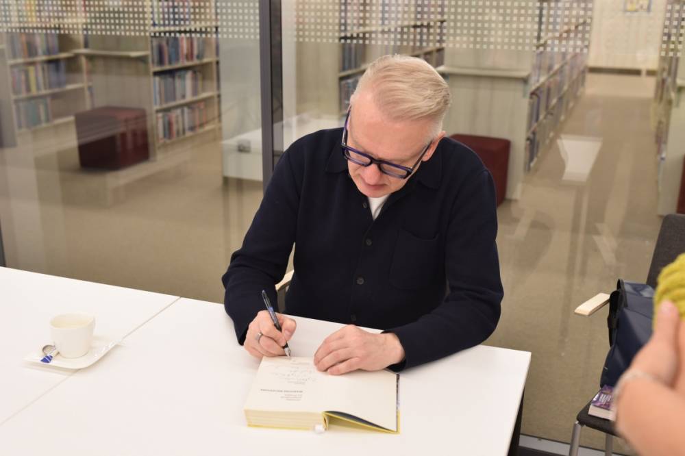 Na zdjęciu widzimy pisarza siedzącego przy białym stole, który z pełnym skupieniem podpisuje książkę. Na stole stoi filiżanka po kawie z łyżeczką na spodku, co sugeruje dłuższą, intensywną pracę podczas spotkania autorskiego. W tle znajduje się szklana ściana, za którą widać regały z książkami w bibliotece. Atmosfera jest spokojna i skupiona, oddając klimat literackiego wydarzenia.