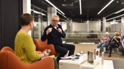 Na zdjęciu widać dwie osoby siedzące na pomarańczowych fotelach na scenie podczas wydarzenia. Osoba po lewej stronie, ubrana w zielony sweter, trzyma mikrofon i zwraca się w stronę rozmówcy. Osoba po prawej stronie, w ciemnym stroju, przemawia do mikrofonu, patrząc na publiczność. Na stoliku przed nimi znajdują się książki oraz szklanki z wodą. W tle widać nowoczesne wnętrze z przeszklonymi balustradami, a po prawej stronie fragment publiczności, która uśmiecha się i z uwagą słucha rozmowy.