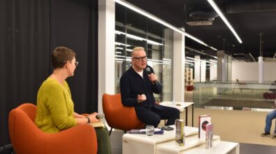 Na zdjęciu znajduje się dwoje ludzi uczestniczących w rozmowie na scenie. Po lewej stronie siedzi osoba w zielonym swetrze i okularach, trzymająca mikrofon i patrząca w stronę swojego rozmówcy. Po prawej stronie siedzi mężczyzna w ciemnym ubraniu, również z mikrofonem w ręce, gestykulujący podczas wypowiedzi. Przed nimi na białym stoliku stoją książki, notes oraz szklanka z wodą. W tle widać nowoczesne wnętrze z przeszklonymi balustradami i dobrze oświetlonymi regałami. Atmosfera sugeruje spotkanie autorskie