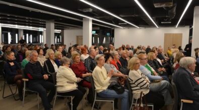 Na zdjęciu widoczna jest duża grupa osób uczestniczących w spotkaniu. Przestrzeń jest jasna, nowoczesna, z charakterystycznym oświetleniem sufitowym w postaci długich, prostokątnych lamp. Uczestnicy siedzą na krzesłach, uważnie słuchając. Widoczna jest różnorodność wiekowa, choć dominują osoby starsze. W tle znajdują się przeszklone drzwi oraz część osób stojących. Atmosfera wydaje się skupiona, co sugeruje udział w wydarzeniu kulturalnym.