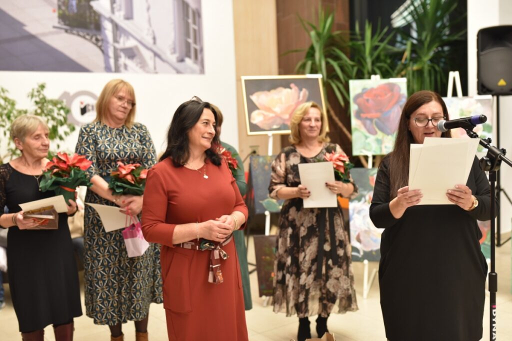 Zdjęcie przedstawia grupę kobiet podczas wernisażu. Jedna z kobiet przemawia do mikrofonu, trzymając kartki z tekstem. Pozostałe kobiety stoją obok, uśmiechając się i trzymając kwiaty oraz dyplomy. W tle widoczne są obrazy z motywami kwiatowymi oraz elementy dekoracyjne wnętrza.