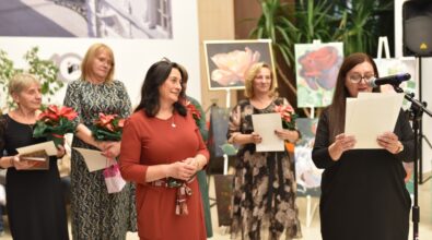 Zdjęcie przedstawia grupę kobiet podczas wernisażu. Jedna z kobiet przemawia do mikrofonu, trzymając kartki z tekstem. Pozostałe kobiety stoją obok, uśmiechając się i trzymając kwiaty oraz dyplomy. W tle widoczne są obrazy z motywami kwiatowymi oraz elementy dekoracyjne wnętrza.
